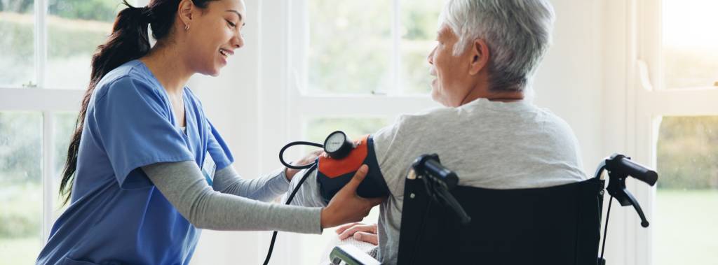Ung kvinnelig helsearbeider måler blodtrykk på eldre dame i rullestol