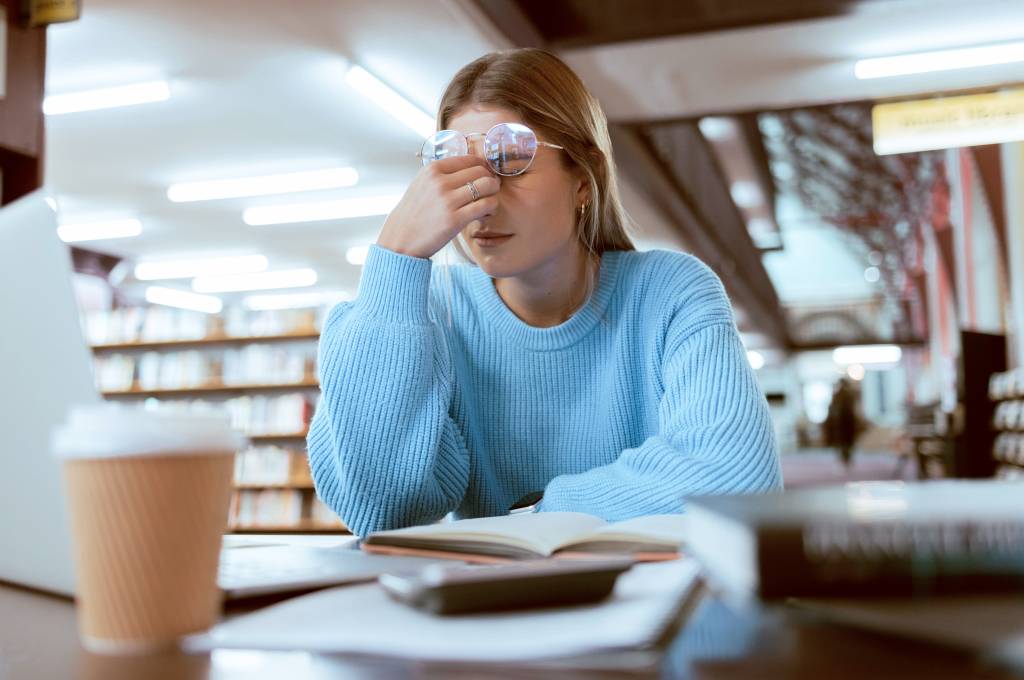 Jente sitter på bibliotek. Tar seg til hodet og ser sliten ut.!''