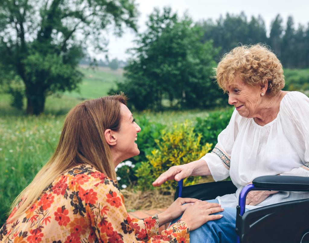 Eldre dame sitter i rullestol med ung jente sittende på huk foran seg