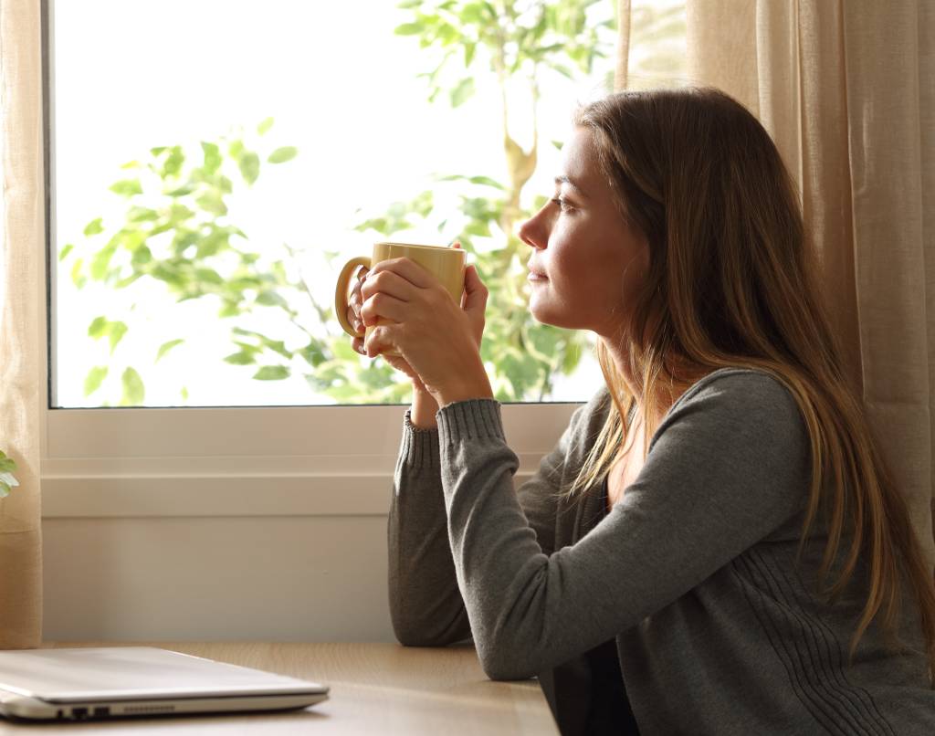 Jente som sitter med en kopp i hendene og ser tankefull ut av vinduet