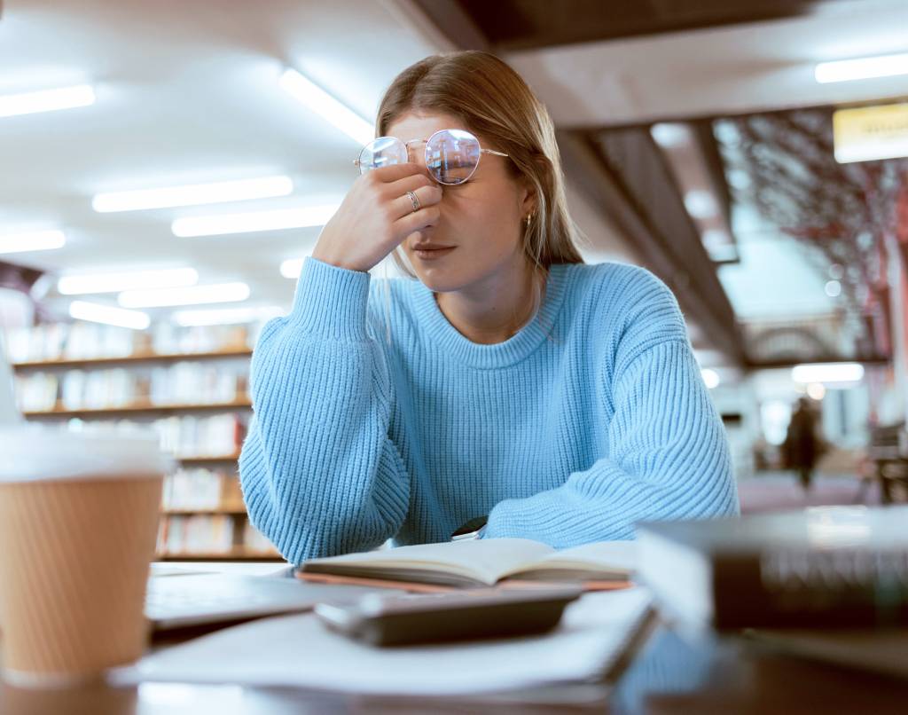 Jente sitter på bibliotek. Tar seg til hodet og ser sliten ut.